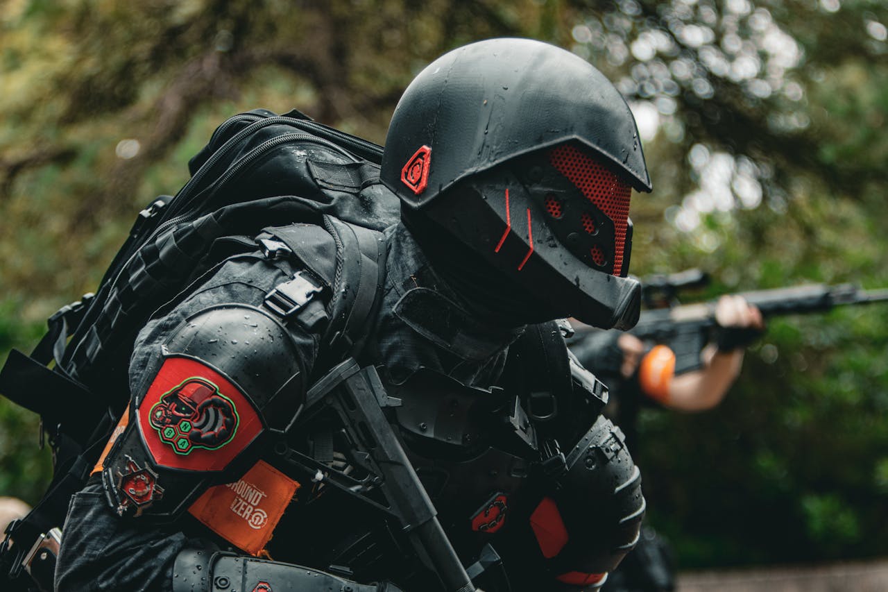 Services A soldier in advanced tactical armor with a firearm crouches in a forested area during a mission.